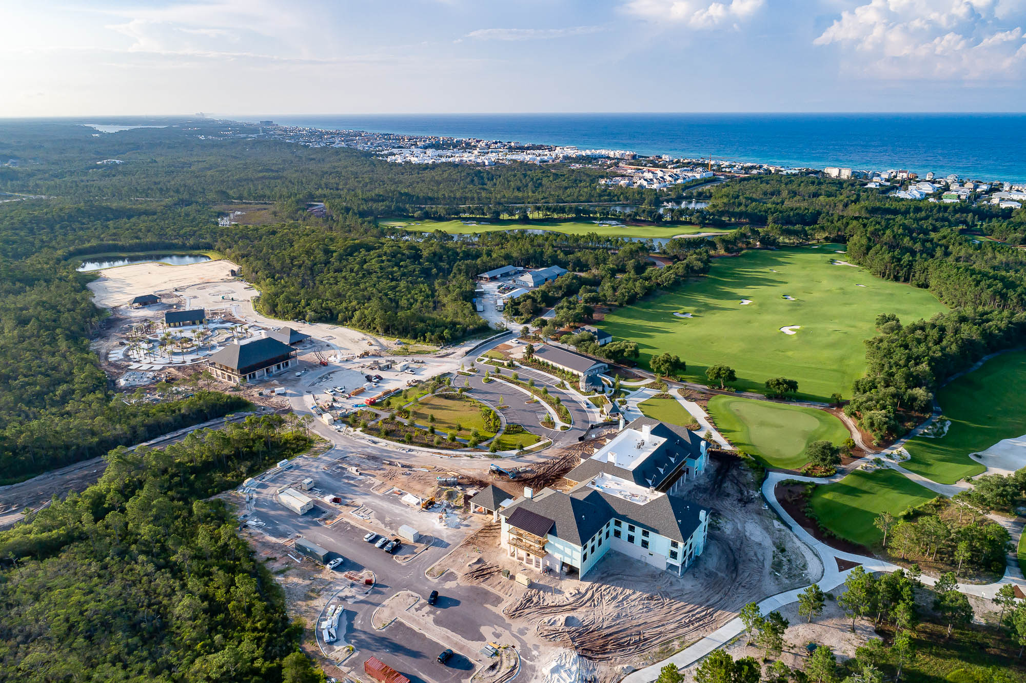 An aerial photo of the new Camp Creek Inn and new Camp Creek amenities construction progress, next to the bright green fairways of Camp Creek Golf Course, and its proximity to the Gulf of Mexico in the background