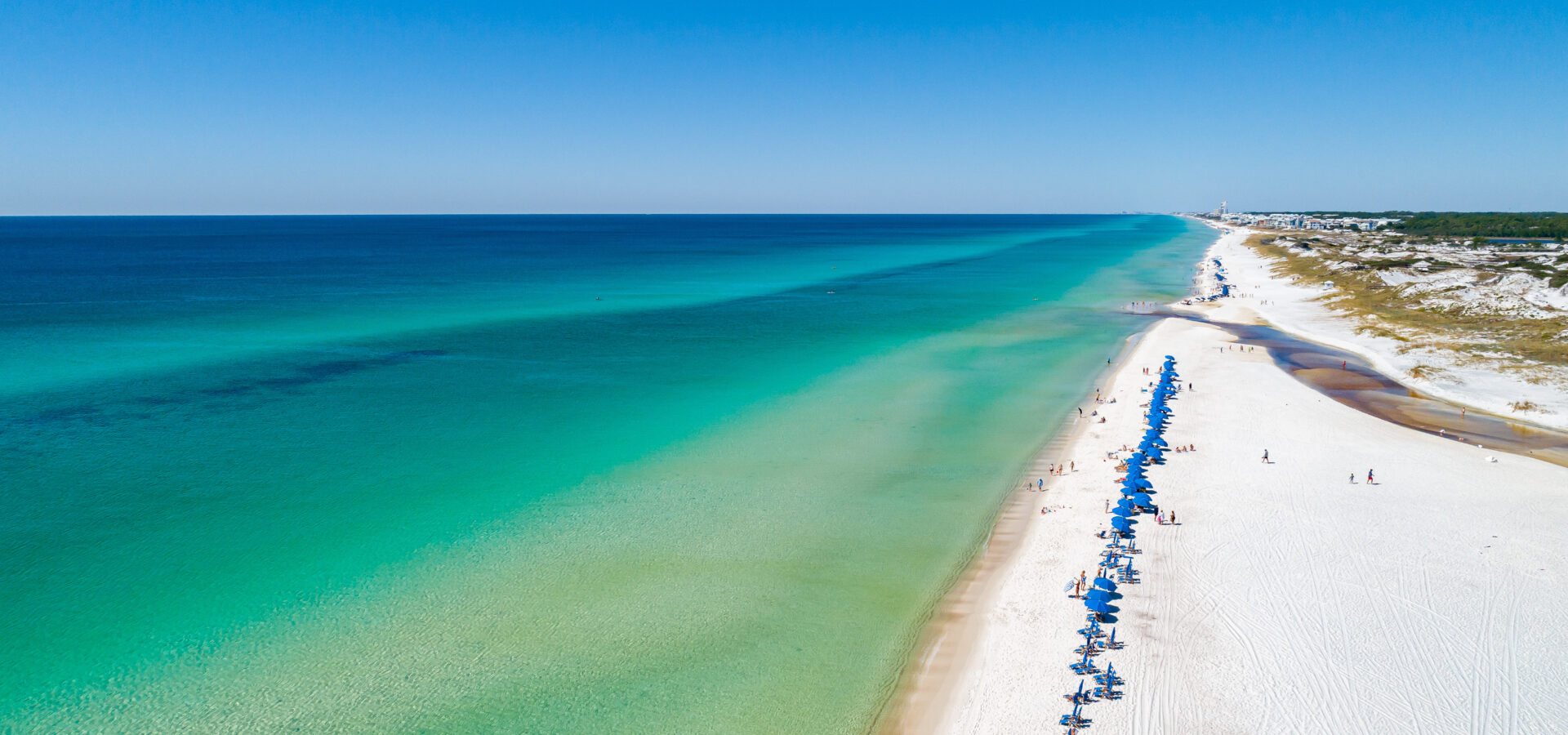 Bright blue beach chairs and umbrellas lined perfectly in a row on the white sandy shoreline of Watersound Beach, overlooking the beautiful emerald green and blue waters of the Gulf of Mexico