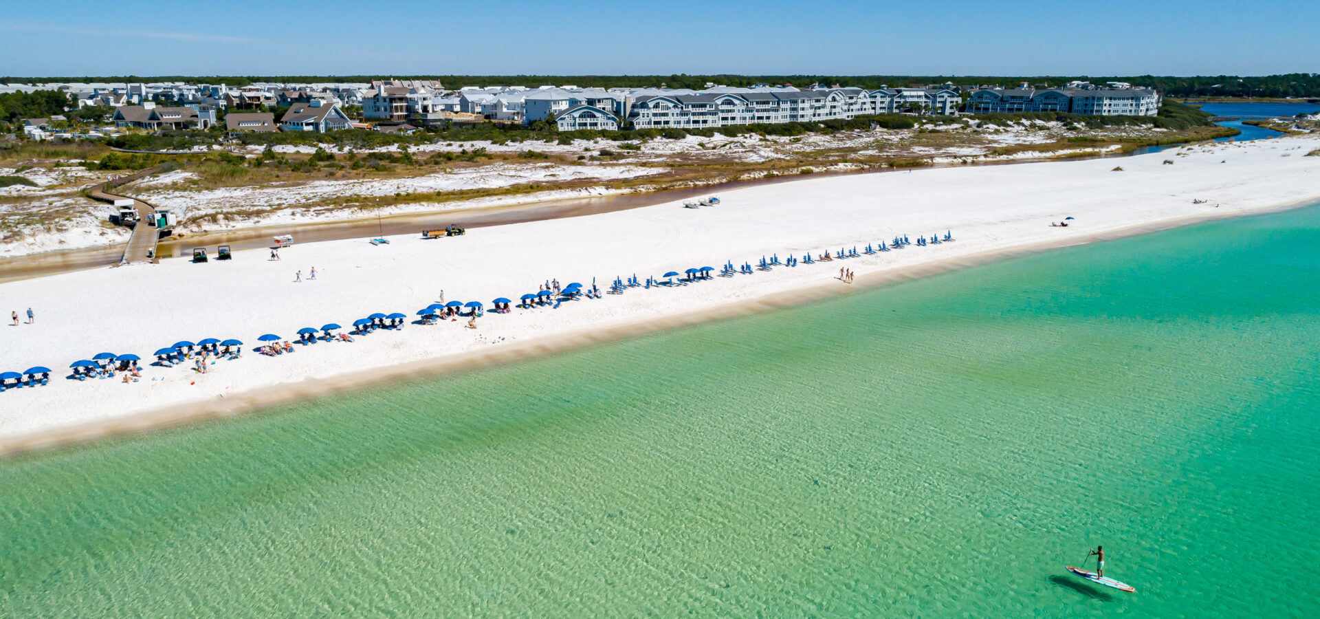 Bright blue beach chairs and umbrellas lined perfectly in a row on the white sandy shoreline of Watersound Beach, overlooking the beautiful emerald green and blue waters of the Gulf of Mexico where a man is paddleboarding on the flat water surface
