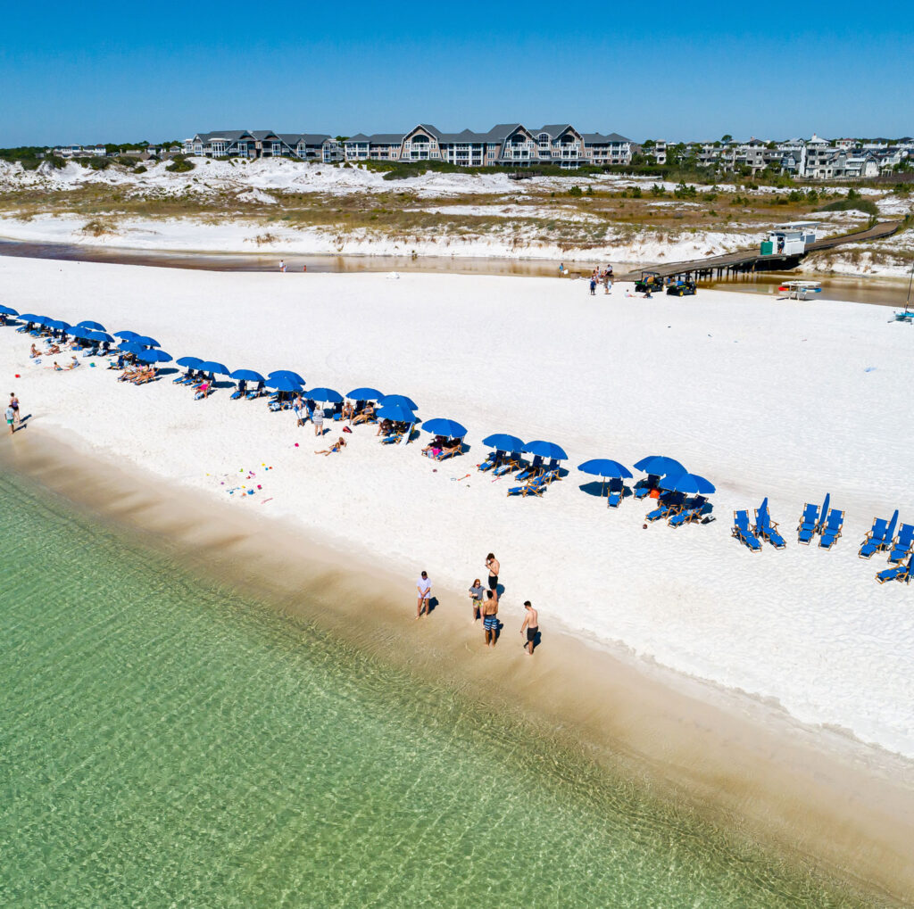 Aerial photo of Watersound Club members enjoying their complimentary blue beach chairs and umbrellas that are dotted along the sugar white sand shoreline looking out at the crystal clear emerald waters of the Gulf of Mexico