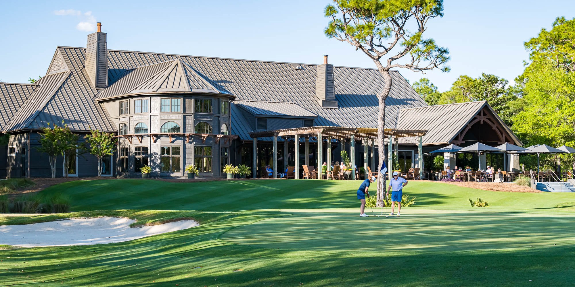 A couple putts on the 18th green at Shark's Tooth Golf Course as others look on while dining on the clubhouse terrace.